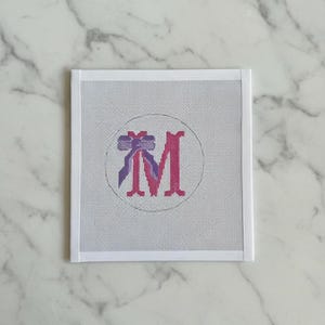 May include: A square needlepoint canvas with a white border. The design features a pink letter "M" inside a circle, with a purple bow at the top. The background is a light grey, and the canvas is on a white marble surface.