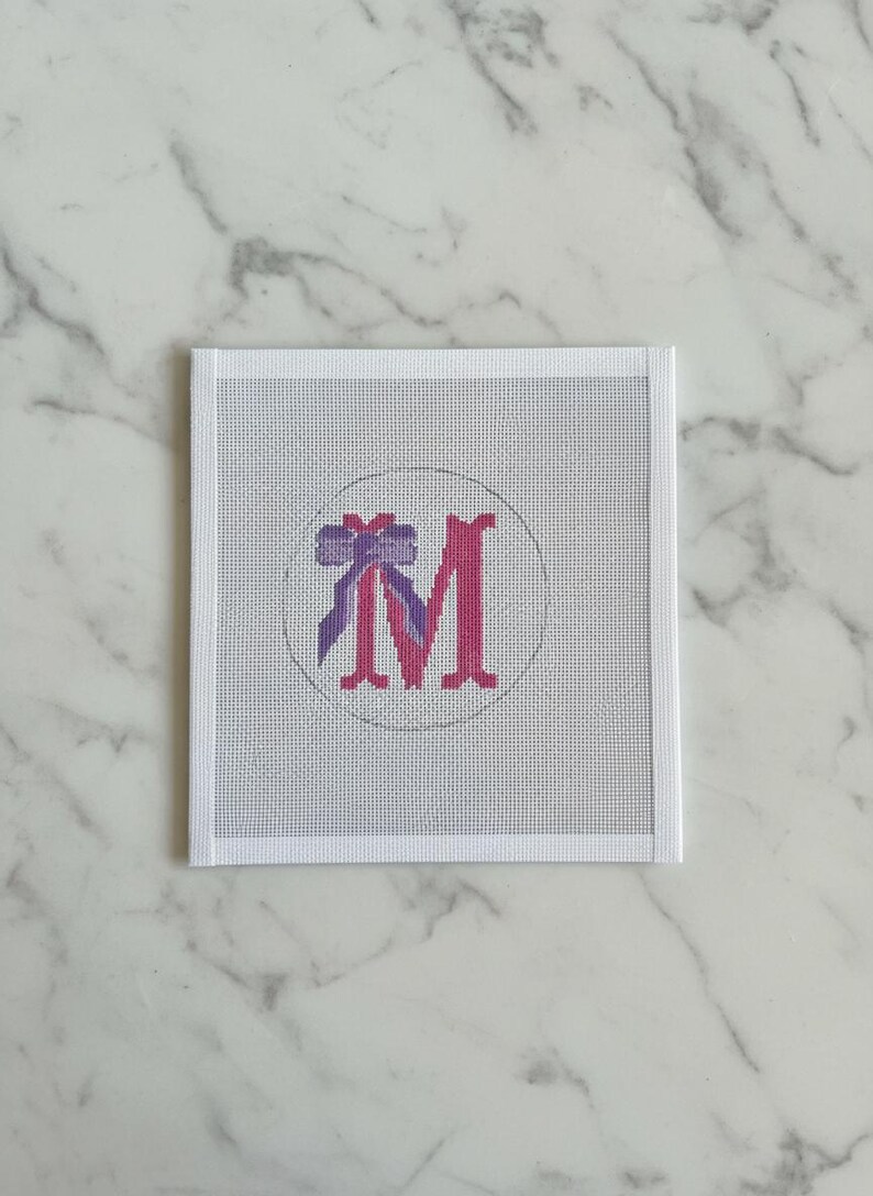 May include: A square needlepoint canvas with a white border. The design features a pink letter "M" inside a circle, with a purple bow at the top. The background is a light grey, and the canvas is on a white marble surface.