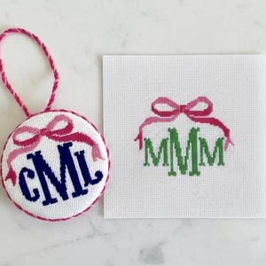 May include: A round, white needlepoint ornament featuring a dark blue monogram CML topped with a pink bow, finished with a pink twisted cord hanger. Beside it is a square white canvas with a green monogram MMM and a pink bow design.