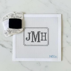 May include: A white needlepoint canvas with the initials "JMH" in a gray frame. A spool of black thread and a white thread spool are next to the canvas. The canvas has the text "NCCo" in the bottom right corner.