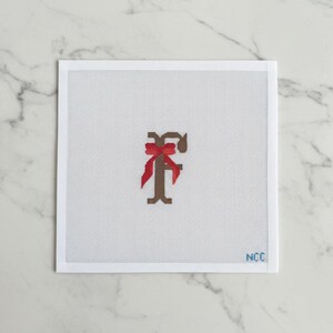 May include: A needlepoint canvas featuring a brown letter "F" adorned with a red bow. The design is centered on a white background with a grid pattern. The lower right corner has the initials "NCC".