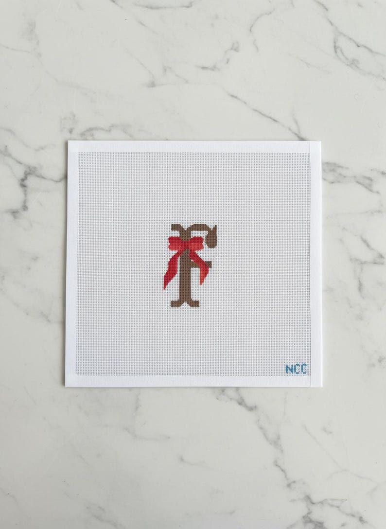 May include: A needlepoint canvas featuring a brown letter "F" adorned with a red bow. The design is centered on a white background with a grid pattern. The lower right corner has the initials "NCC".