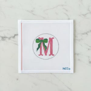 May include: White needlepoint canvas with a pink letter "M" inside a gray circle, adorned with a green bow. A thin red line runs along the left edge. The text "NCCo" is in the bottom right corner.