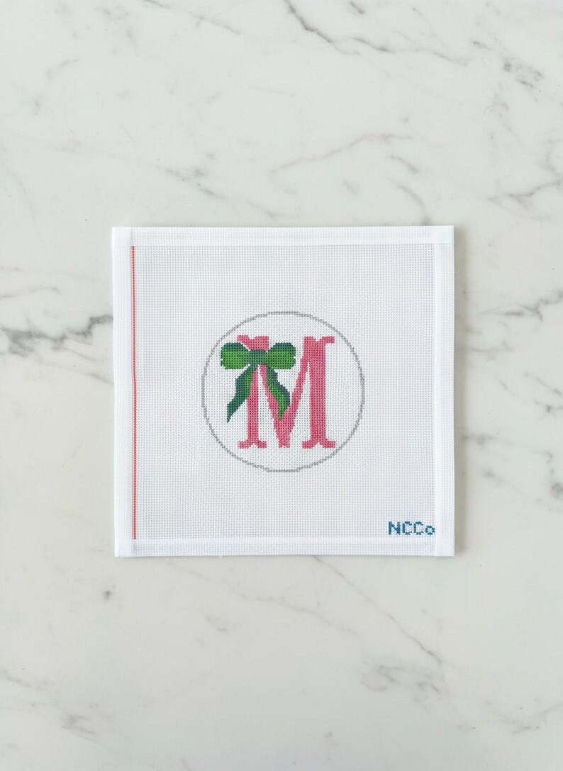 May include: White needlepoint canvas with a pink letter "M" inside a gray circle, adorned with a green bow. A thin red line runs along the left edge. The text "NCCo" is in the bottom right corner.