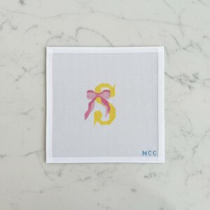 May include: Needlepoint canvas featuring a yellow letter "S" with a pink bow. The letter is centered on a white canvas with a grid pattern. The lower right corner has the initials "NCC".