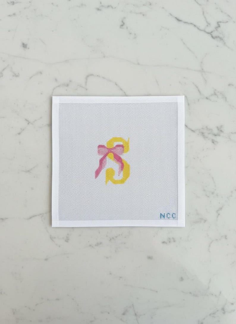 May include: Needlepoint canvas featuring a yellow letter "S" with a pink bow. The letter is centered on a white canvas with a grid pattern. The lower right corner has the initials "NCC".