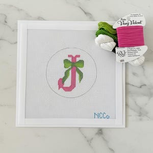 May include: Needlepoint canvas with a pink letter "J" and a green bow design inside a circle. The canvas is framed in white and includes the text "NCCo" in blue. A spool of pink, green, and white thread sits to the side.