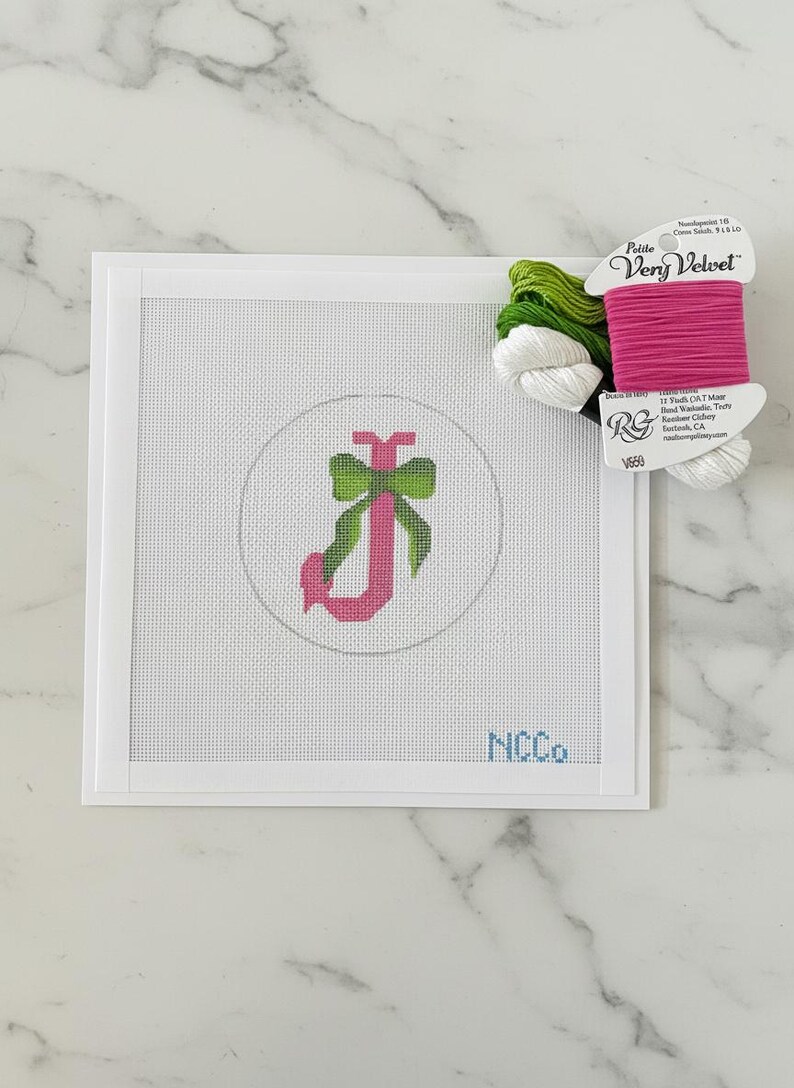 May include: Needlepoint canvas with a pink letter "J" and a green bow design inside a circle. The canvas is framed in white and includes the text "NCCo" in blue. A spool of pink, green, and white thread sits to the side.