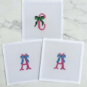 May include: Three white needlepoint canvases with embroidered letters and bows. One canvas features a pink "C" with a green bow. The other two canvases display a pink "A" with a blue bow.