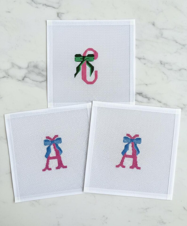 May include: Three white needlepoint canvases with embroidered letters and bows. One canvas features a pink "C" with a green bow. The other two canvases display a pink "A" with a blue bow.
