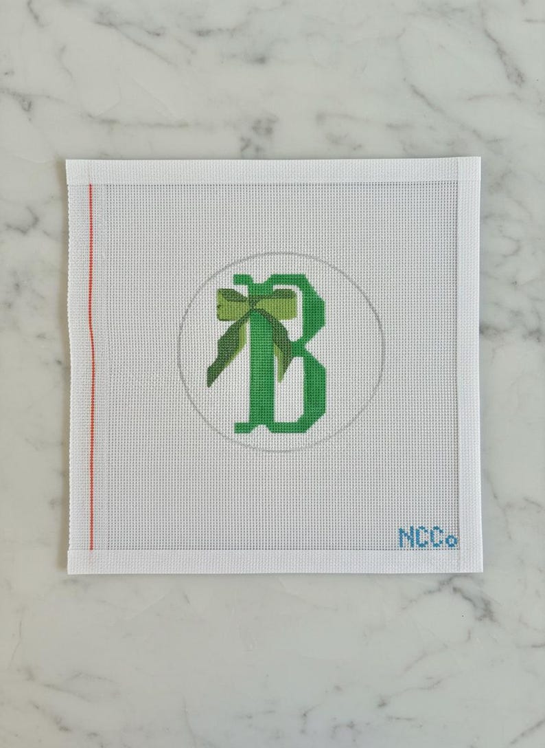 May include: Needlepoint canvas featuring a green letter "B" with a bow design inside a gray circle. The canvas is white with a red stripe on the left edge and the letters "NCCo" in the bottom right corner.