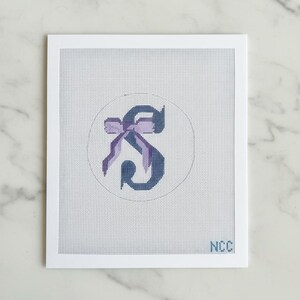 May include: A needlepoint canvas featuring the letter "S" in a dark blue hue, adorned with a purple bow. The design is set within a white circle on a white background, framed by a white border. The initials "NCC" are visible in the lower right corner.