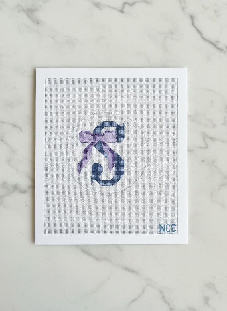 May include: A needlepoint canvas featuring the letter "S" in a dark blue hue, adorned with a purple bow. The design is set within a white circle on a white background, framed by a white border. The initials "NCC" are visible in the lower right corner.
