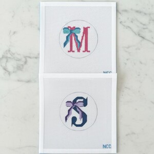 May include: Two needlepoint canvases with embroidered letters and bows. The top canvas features a pink "M" with a teal and blue bow. The bottom canvas displays a dark blue "S" with a purple bow. Both are set within a white circle.