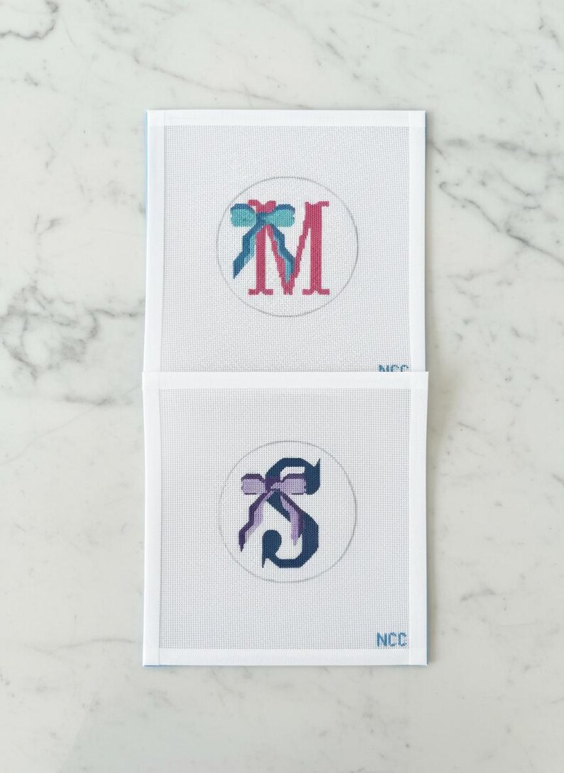 May include: Two needlepoint canvases with embroidered letters and bows. The top canvas features a pink "M" with a teal and blue bow. The bottom canvas displays a dark blue "S" with a purple bow. Both are set within a white circle.
