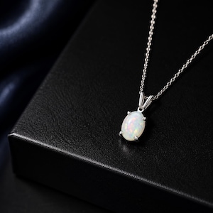 Puede incluir: Un collar de plata con un colgante de ópalo ovalado. El ópalo es de color blanco lechoso con sutiles destellos de color. El colgante está engastado en un engaste de cuatro garras y cuelga de una delicada cadena. El collar se muestra sobre una superficie negra.