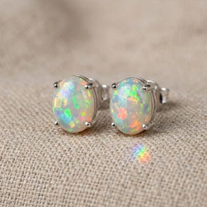 Könnte beinhalten: Ein Paar ovale Opal-Ohrstecker. Die Opale zeigen ein Farbspiel mit grünen, orangefarbenen und blauen Blitzen. Die Opale sind in einer silberfarbenen Metalleinfassung gefasst. Die Ohrringe werden auf einem neutralfarbenen Stoff präsentiert.