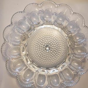 Pode incluir: Prato de servir de vidro transparente com borda ondulada e área central texturizada. O prato apresenta reentrâncias moldadas em forma de ovo ao redor do perímetro, perfeitas para servir aperitivos. O vidro tem uma aparência delicada e vintage.