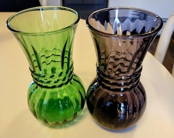 Op de afbeelding: Twee glazen vaasjes voor boeketten, een groene en een paarse, met een geribbeld ontwerp. De vaasjes staan op een wit oppervlak.