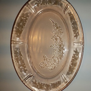 May include: Oval pink glass ashtray with floral detailing. The ashtray has a raised floral pattern around the edge and a central floral design. It includes four cigarette rests.