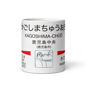 Può includere: Una tazza in ceramica bianca con testo nero che recita KAGOSHIMA-CHŪŌ, caratteri giapponesi e un'illustrazione grafica di una persona davanti a una montagna. Il design include barre orizzontali rosse e testo aggiuntivo in giapponese e inglese.