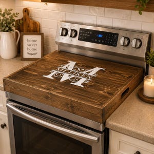 May include: A dark stained wooden stove cover with a white monogram and the name "Milliken" printed on it. The cover is rectangular with handles and sits on top of a stainless steel stove.
