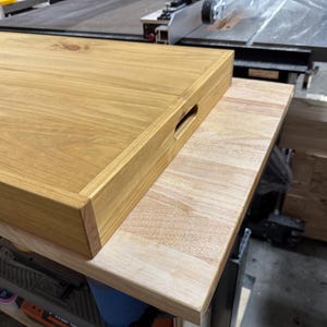 May include: A rectangular wooden tray with a handle cut-out, resting on a light-colored wooden surface. The tray is made of light brown wood with visible wood grain. The tray is approximately 18 inches long and 12 inches wide.