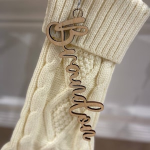 May include: A wooden ornament with the name "Brandon" in a cursive font hangs from a silver wire. The ornament is suspended in front of a cream-colored cable-knit Christmas stocking. The background is blurred.