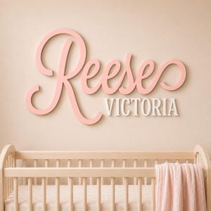 May include: A wall-mounted sign featuring the name Reese in large, cursive, light pink lettering, positioned above the name VICTORIA in smaller, white, block-style capital letters, displayed above a light-colored wooden crib with a pink blanket.
