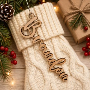 May include: A wooden ornament with the name "Brandon" in a cursive font hangs from a silver hook. The ornament is placed on a cream-colored cable-knit Christmas stocking. A pine cone, berries, and a wrapped gift are in the background.