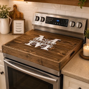 May include: A dark stained wooden stove top cover with a white monogram and the name "Milliken" printed on it. The cover is rectangular with handles and sits on a stainless steel stove.