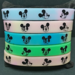 Puede incluir: Una pila de cinco pulseras de silicona de colores. Cada pulsera es de un color pastel diferente, incluyendo azul, rosa, verde y amarillo. Las pulseras est&aacute;n decoradas con un patr&oacute;n repetido de caras negras de Mickey Mouse.