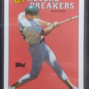 May include: A baseball card featuring Mark McGwire in action, swinging a bat. The card has a red background with the text "'87 RECORD BREAKERS" and the Topps logo. McGwire is wearing a green helmet and white uniform.