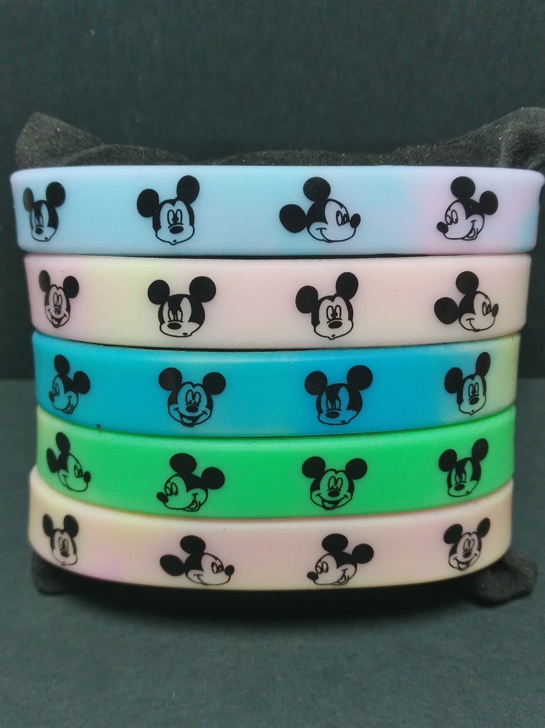 Puede incluir: Una pila de cinco pulseras de silicona de colores. Cada pulsera es de un color pastel diferente, incluyendo azul, rosa, verde y amarillo. Las pulseras est&aacute;n decoradas con un patr&oacute;n repetido de caras negras de Mickey Mouse.