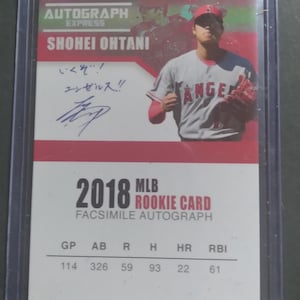 May include: A 2018 Shohei Ohtani MLB rookie card in a protective plastic sleeve. The card features a photograph of the player in a grey Angels uniform, a facsimile autograph with Japanese text, and a table of 2018 season statistics including GP, AB, R, H, HR, and RBI.