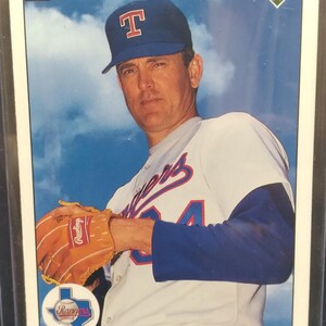 May include: A baseball card featuring Nolan Ryan in a Texas Rangers uniform. The card shows Ryan wearing a blue cap with a red "T" and holding a baseball glove. The Rangers logo is visible. The card is from the 1990 Upper Deck series.