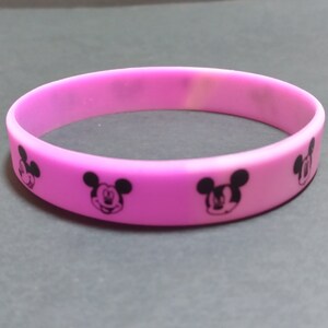 Puede incluir: Una pulsera de silicona rosa con siluetas negras de Mickey Mouse impresas en ella.