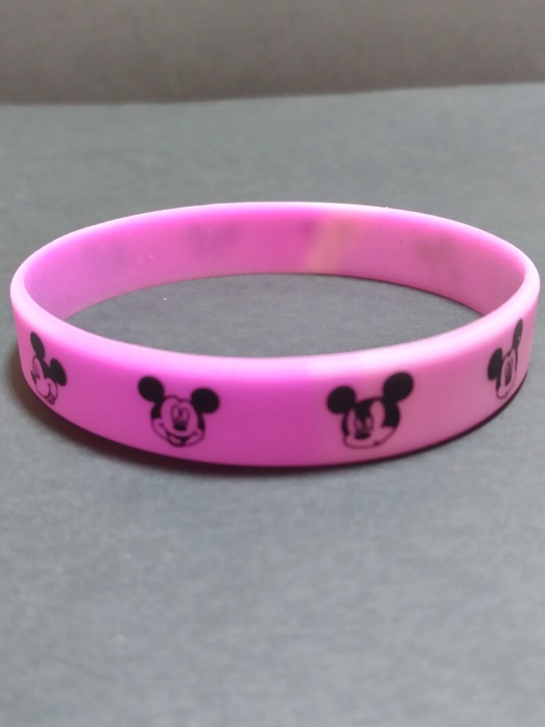 Puede incluir: Una pulsera de silicona rosa con siluetas negras de Mickey Mouse impresas en ella.