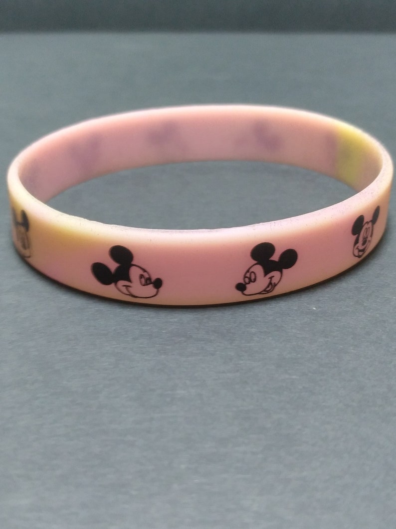 Puede incluir: Una pulsera de goma rosa y amarilla con dise&ntilde;os negros de Mickey Mouse. La pulsera es una banda s&oacute;lida con la cara del personaje de dibujos animados impresa alrededor de la circunferencia. El fondo es gris liso.