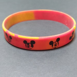 Puede incluir: Una pulsera de silicona roja y amarilla con un patr&oacute;n repetido de cabezas de Mickey Mouse negras.