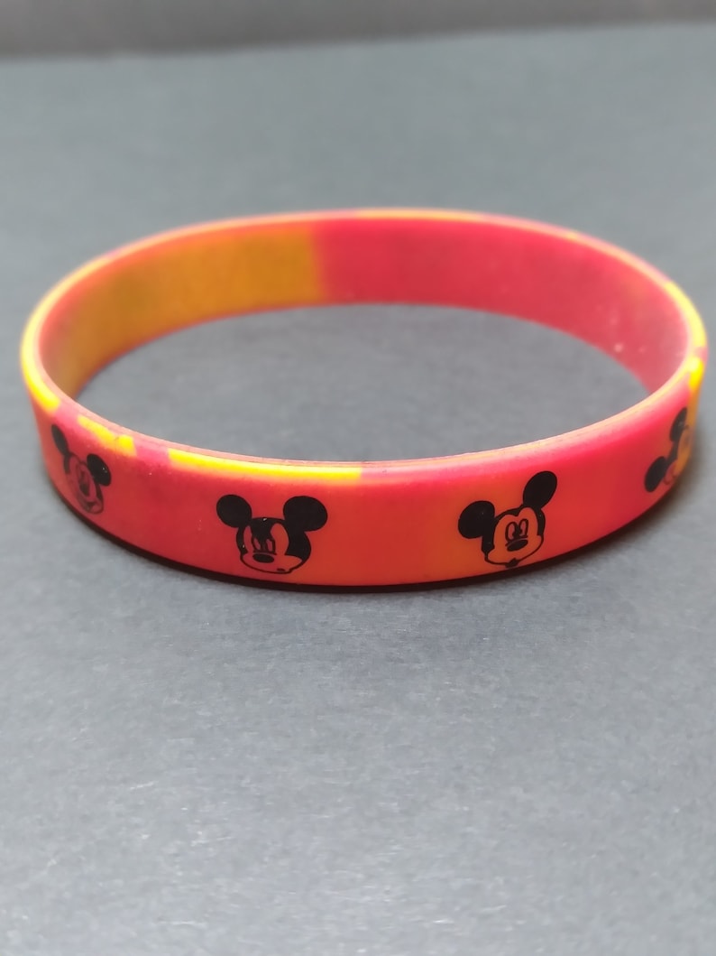 Puede incluir: Una pulsera de silicona roja y amarilla con un patr&oacute;n repetido de cabezas de Mickey Mouse negras.