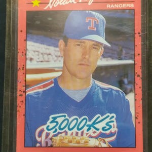 May include: A 1990 Donruss baseball card featuring Nolan Ryan of the Texas Rangers. The card has a red border and displays Ryan in a blue Rangers uniform and cap. The card also features the text "5,000 K's".