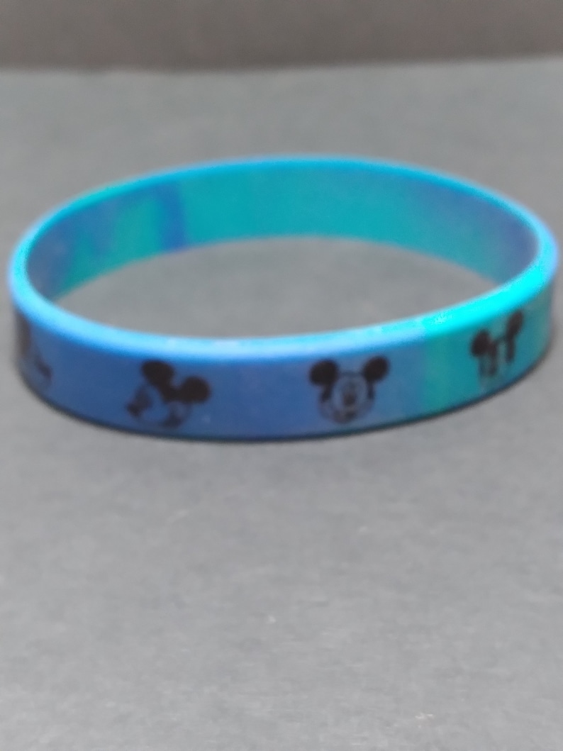 Puede incluir: Una pulsera de silicona azul con un patr&oacute;n repetido de cabezas de Mickey Mouse negras.