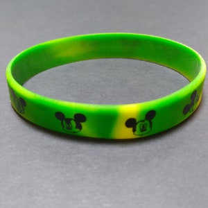 Puede incluir: Una pulsera de silicona verde y amarilla con un dise&ntilde;o de Mickey Mouse negro.