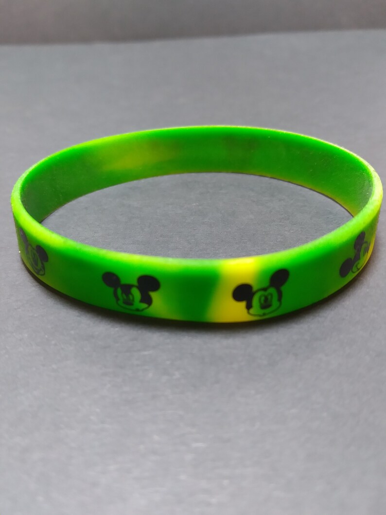 Puede incluir: Una pulsera de silicona verde y amarilla con un dise&ntilde;o de Mickey Mouse negro.