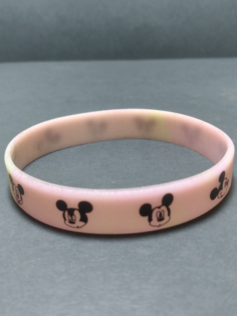 Puede incluir: Una pulsera de silicona rosa claro con caras negras de Mickey Mouse. La pulsera es una banda s&oacute;lida, con la cara del personaje de dibujos animados repiti&eacute;ndose alrededor de la circunferencia. Un accesorio sencillo e informal.