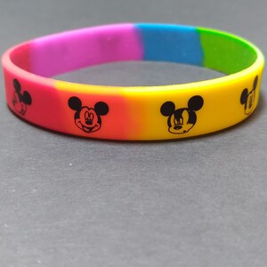 Puede incluir: Una pulsera de silicona colorida con un patr&oacute;n de arco&iacute;ris y cuatro siluetas negras de Mickey Mouse.