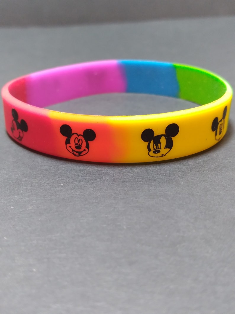 Puede incluir: Una pulsera de silicona colorida con un patr&oacute;n de arco&iacute;ris y cuatro siluetas negras de Mickey Mouse.