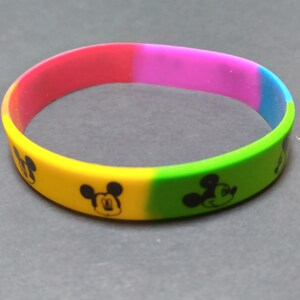 Puede incluir: Una pulsera de silicona multicolor con un patr&oacute;n repetido de la cabeza de Mickey Mouse. La pulsera es roja, rosa, amarilla, verde y azul.