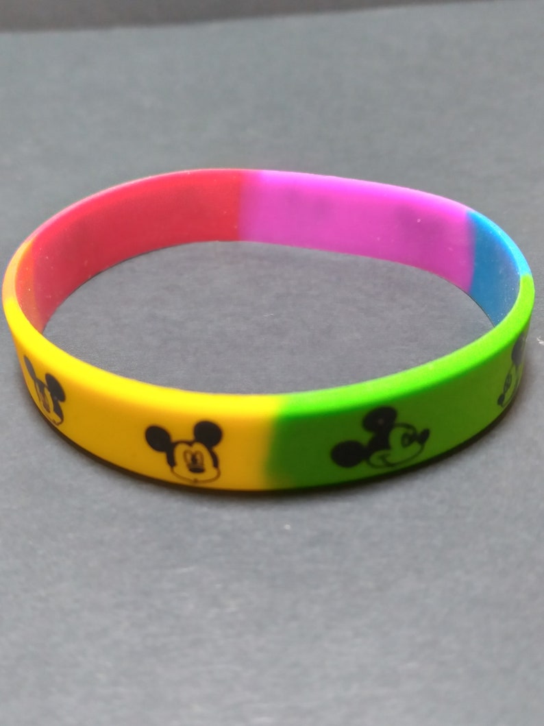 Puede incluir: Una pulsera de silicona multicolor con un patr&oacute;n repetido de la cabeza de Mickey Mouse. La pulsera es roja, rosa, amarilla, verde y azul.